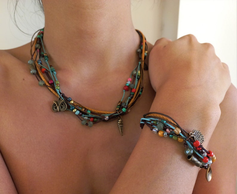 Op de afbeelding: Een multi-strand ketting- en armbandset. De ketting en armband hebben bruine, bruine en blauwe koorden versierd met kleurrijke kralen in rood, groen en geel. Beide stukken hebben metalen bedels, waaronder een uil en een punt.
