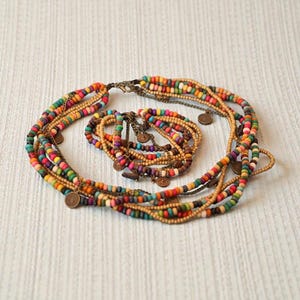 May include: A multi-strand necklace and bracelet set featuring colorful wooden beads in shades of orange, green, yellow, and purple. The necklace has small, round, bronze-colored coin charms. The jewelry is arranged on a white textured surface.