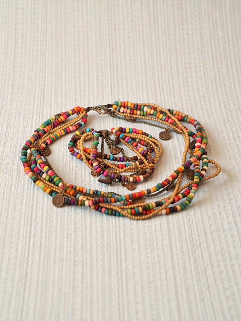 May include: A multi-strand necklace and bracelet set featuring colorful wooden beads in shades of orange, green, yellow, and purple. The necklace has small, round, bronze-colored coin charms. The jewelry is arranged on a white textured surface.