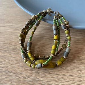 May include: A multi-strand bracelet featuring olive green, yellow, and gray beads. The bracelet has a mix of small round beads and larger rectangular and oval beads. The clasp is gold-toned.