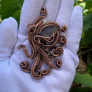 May include: A wire-wrapped copper pendant shaped like an octopus, featuring a central oval labradorite gemstone, small dark red garnet beads, and a small dark blue starfish accent. The intricate metalwork consists of coiled and woven copper wire.