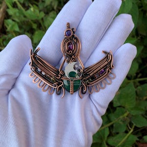 May include: A copper-colored pendant with an ornate design, featuring purple and green gemstones. The pendant has a central green stone and a logo that reads "Indonesia Stone Artworks". The wings are detailed with small, curved elements.