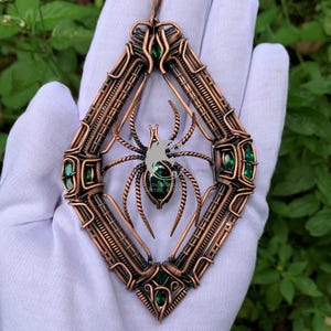 以下が含まれることがあります： ダイヤモンド型のフレームとワイヤーラップデザインが特徴の銅色のペンダント。ペンダントの中央にはクモのデザインがあり、緑色の宝石がアクセントになっています。ペンダントは白い背景に保持されています。
