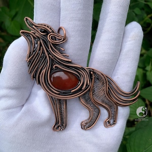 Puede incluir: Colgante de alambre de color cobre con forma de lobo aullando. El colgante presenta una piedra de color marrón rojizo en el centro y se sostiene en una mano enguantada de blanco. El lobo está detallado con intrincados trabajos de alambre, mostrando la artesanía.