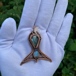May include: A wire-wrapped copper pendant featuring a teardrop-shaped labradorite stone at the top and a smaller, round, light blue opal-like cabochon at the base. The metal wire is intricately coiled and shaped into a symmetrical, tail-like design.
