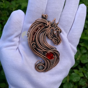May include: Copper-toned pendant in the shape of a horse's head, featuring intricate wirework and a red gemstone accent. The pendant is suspended from a loop at the top and has a swirling design at the bottom. The horse's head is detailed with a mane and eye.