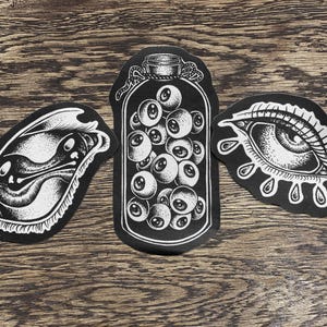 May include: Three black and white embroidered patches featuring graphic illustrations. The left patch shows a stylised eye, the centre patch depicts a jar filled with multiple eyeballs, and the right patch displays an eye with teardrop shapes below it.