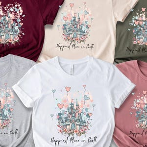 May include: A collection of six t-shirts in various colors, including burgundy, beige, olive green, gray, white, and mauve. Each shirt features a castle design with floral and heart accents, and the text "Happiest Place on Earth".