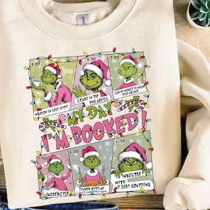 May include: A beige crewneck sweatshirt featuring a graphic print of a green character in a Santa hat. The design includes six panels showing daily activities with times and the text: MY DAY I'M BOOKED! surrounded by colourful Christmas light bulbs.