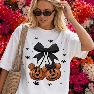 May include: A white short-sleeved t-shirt featuring a graphic of two orange jack-o'-lanterns with mouse-shaped ears, connected by a large black bow. The design includes small black bat silhouettes and stars scattered around the pumpkins.