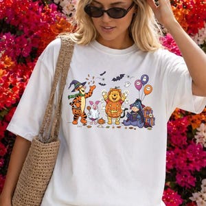 May include: A white short-sleeve t-shirt featuring a graphic print of Tigger, Piglet, Winnie the Pooh, and Eeyore in Halloween costumes. The characters are surrounded by small bats, ghosts, and balloons. A tan woven tote bag hangs from the shoulder.