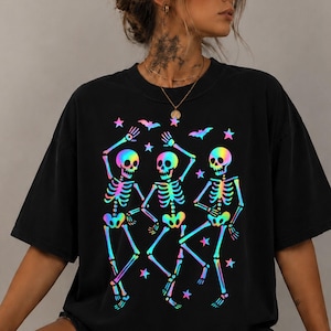 May include: A black short-sleeved t-shirt featuring a graphic print of three dancing skeletons. The skeletons are rendered in a vibrant, iridescent rainbow gradient. The design also includes small stars and bats scattered around the skeletons.
