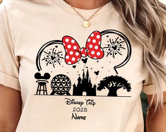 Puede incluir: Una camiseta color crema con un gráfico negro de una silueta de orejas de Mickey rellena de fuegos artificiales, un lazo rojo de lunares blancos y monumentos icónicos. Debajo del gráfico, el texto dice Disney Trip 2025 y Name.
