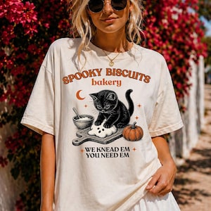 May include: An off-white short-sleeve t-shirt featuring a graphic of a black kitten kneading dough on a wooden board. The text reads SPOOKY BISCUITS bakery and WE KNEAD EM YOU NEED EM. A small bowl, a crescent moon, and a pumpkin are also illustrated.