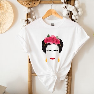 May include: White t-shirt with a black and white illustration of Frida Kahlo wearing a pink flower crown and gold earrings.