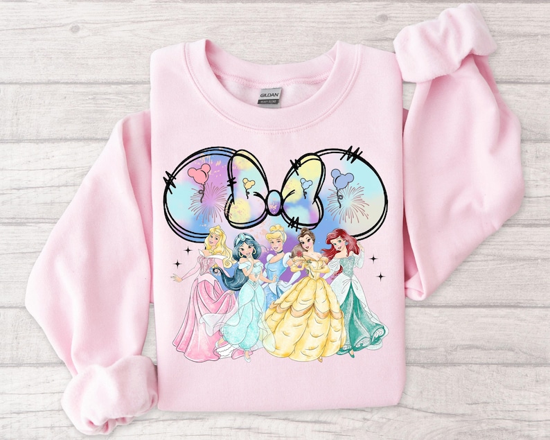 May include: Pink sweatshirt with a tie-dye bow and a group of Disney princesses in front of it. The princesses are Cinderella, Jasmine, Ariel, Belle, and Aurora.