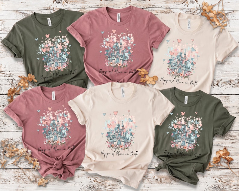 May include: A collection of six short-sleeved t-shirts in various colors, including olive green, dusty rose, and cream. Each shirt features a castle graphic with floral and heart accents, and the text "Happiest Place on Earth".