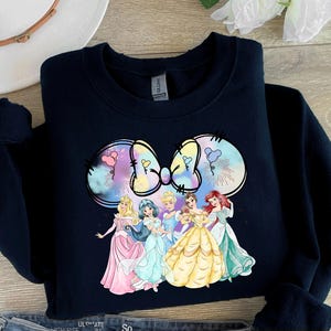 May include: A black sweatshirt with a colorful graphic of a bow and six Disney princesses. The princesses are wearing their signature dresses and are arranged in a semi-circle around the bow.