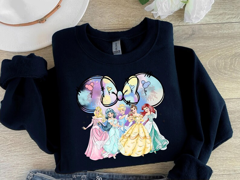 May include: A black sweatshirt with a colorful graphic of a bow and six Disney princesses. The princesses are wearing their signature dresses and are arranged in a semi-circle around the bow.