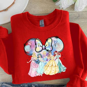 May include: Red sweatshirt with a colorful graphic of a bow with Disney princesses. The princesses are wearing their signature dresses and are arranged in a semi-circle around the bow.