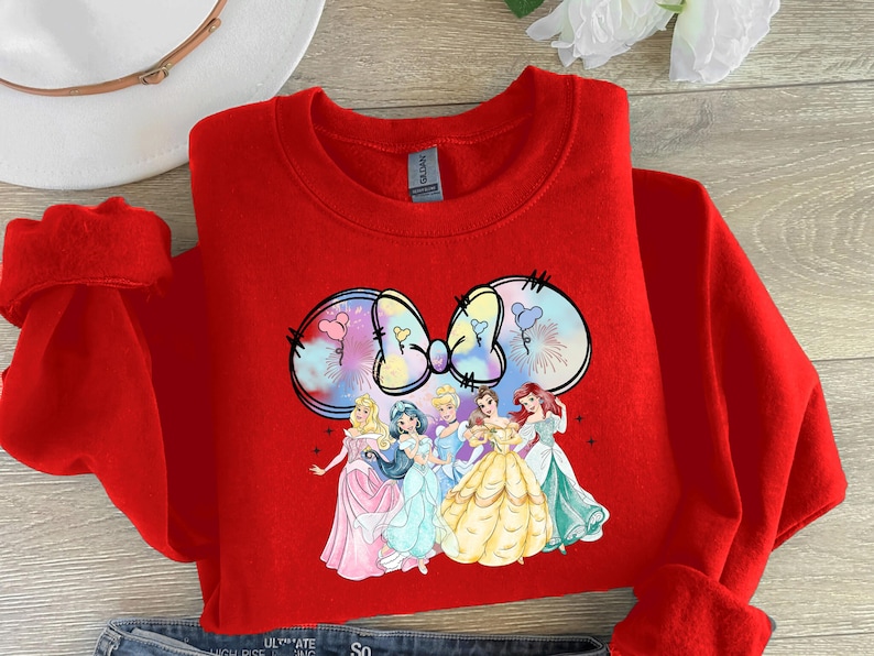 May include: Red sweatshirt with a colorful graphic of a bow with Disney princesses. The princesses are wearing their signature dresses and are arranged in a semi-circle around the bow.