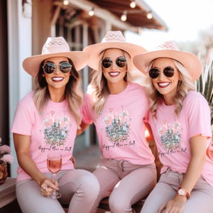 May include: Three people wearing pink t-shirts with a castle graphic and the text "Happiest Place on Earth." They are also wearing sunglasses and pink hats. The image has a soft, warm color palette.