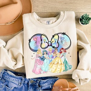 May include: A cream-colored sweatshirt with a colorful tie-dye bow and five Disney princesses in front of it. The princesses are wearing their signature dresses and are arranged in a line. The sweatshirt has a crew neck and long sleeves.