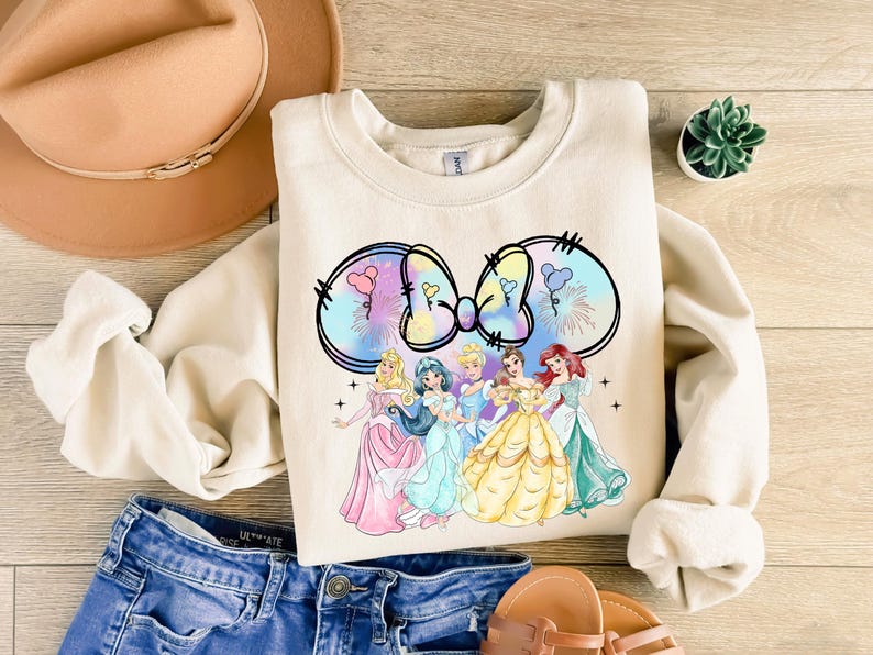 May include: A cream-colored sweatshirt with a colorful tie-dye bow and five Disney princesses in front of it. The princesses are wearing their signature dresses and are arranged in a line. The sweatshirt has a crew neck and long sleeves.