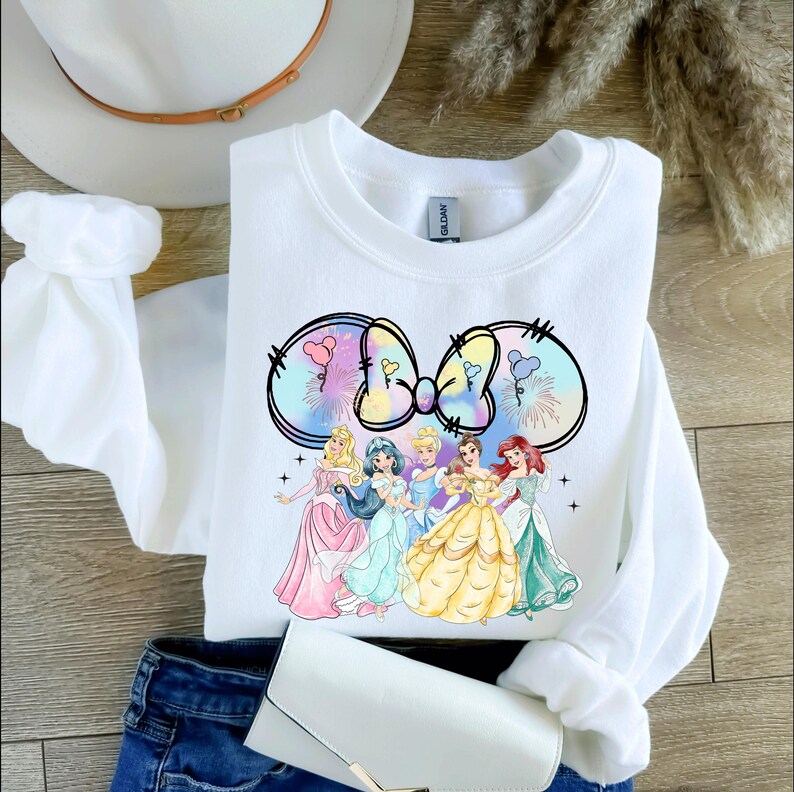 May include: White sweatshirt with a tie-dye bow design featuring Disney princesses. The princesses are wearing their signature dresses and are arranged in a circle around the bow. The bow is in shades of pink, blue, and yellow.