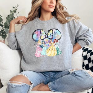 May include: A gray sweatshirt with a graphic of Disney princesses in a Mickey Mouse ear design. The princesses are wearing their signature dresses and are surrounded by a pastel-colored background.
