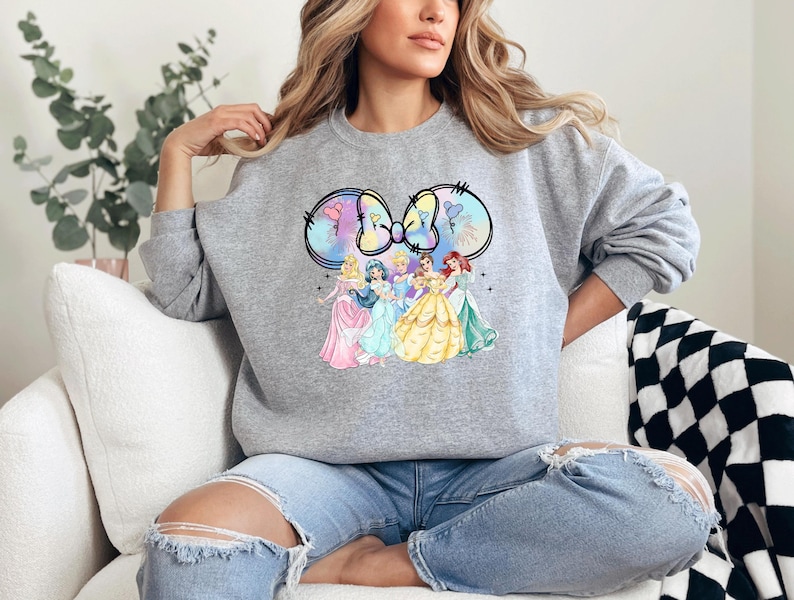 May include: A gray sweatshirt with a graphic of Disney princesses in a Mickey Mouse ear design. The princesses are wearing their signature dresses and are surrounded by a pastel-colored background.