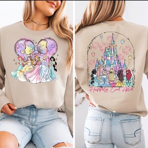 Puede incluir: Una sudadera de color beige con un gr&aacute;fico de princesas Disney frente a un castillo con fuegos artificiales. La parte posterior de la sudadera tiene un gr&aacute;fico del castillo de Cenicienta con fuegos artificiales y el texto "Happily Ever After".