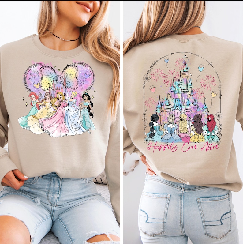 Puede incluir: Una sudadera de color beige con un gr&aacute;fico de princesas Disney frente a un castillo con fuegos artificiales. La parte posterior de la sudadera tiene un gr&aacute;fico del castillo de Cenicienta con fuegos artificiales y el texto "Happily Ever After".