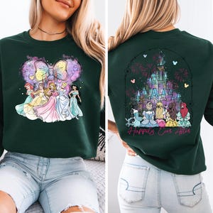 Puede incluir: Una sudadera verde oscuro con una ilustraci&oacute;n colorida de princesas Disney frente a un lazo rosa y un castillo azul claro con fuegos artificiales en la espalda. El texto "Happily Ever After" est&aacute; impreso en la parte posterior de la sudadera.