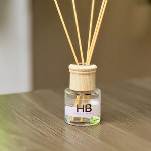 Puede incluir: Un pequeño difusor de aromas de vidrio transparente con una tapa de madera y cañas. La botella tiene una etiqueta blanca con las letras "HB" y las palabras "HINONEY AROMAS". Las cañas son de color marrón claro y se extienden hacia arriba.