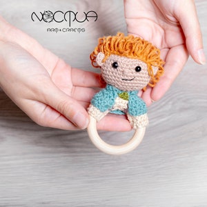 May include: A handmade crochet baby rattle in the shape of a doll with orange hair and a blue jacket. The doll is attached to a wooden ring. The rattle is held in two hands. The brand name "nocpua arm & craems" is in the upper left corner.