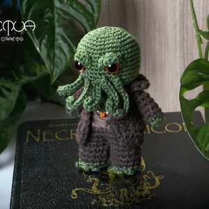 Pode incluir: Brinquedo de pel&uacute;cia Cthulhu verde em croch&ecirc; com olhos vermelhos, vestindo um terno marrom em croch&ecirc;, sentado em um livro preto com letras douradas que dizem "Necronomicon".