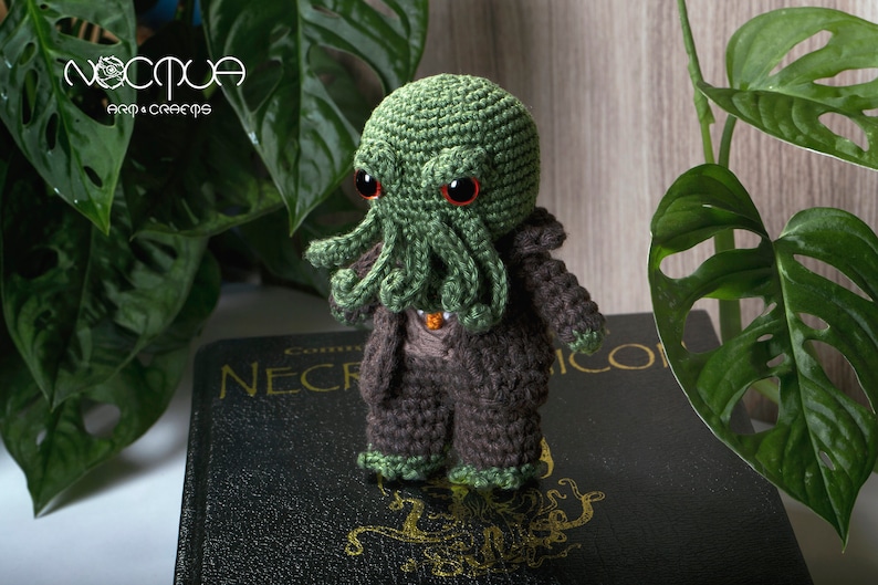 Pode incluir: Brinquedo de pel&uacute;cia Cthulhu verde em croch&ecirc; com olhos vermelhos, vestindo um terno marrom em croch&ecirc;, sentado em um livro preto com letras douradas que dizem "Necronomicon".