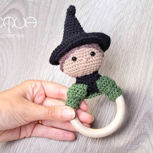 Puede incluir: Una muñeca de bruja de crochet con un sombrero negro y ropa verde y negra unida a un anillo de madera. La muñeca tiene pelo de lana marrón y ojos negros.