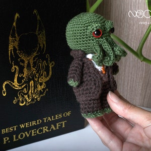 Pode incluir: Um brinquedo de pel&uacute;cia verde e marrom de croch&ecirc; em forma de monstro Cthulhu vestindo um terno marrom. O brinquedo est&aacute; sendo segurado em uma m&atilde;o. O fundo &eacute; um livro com o t&iacute;tulo "The Best Weird Tales of H.P. Lovecraft".