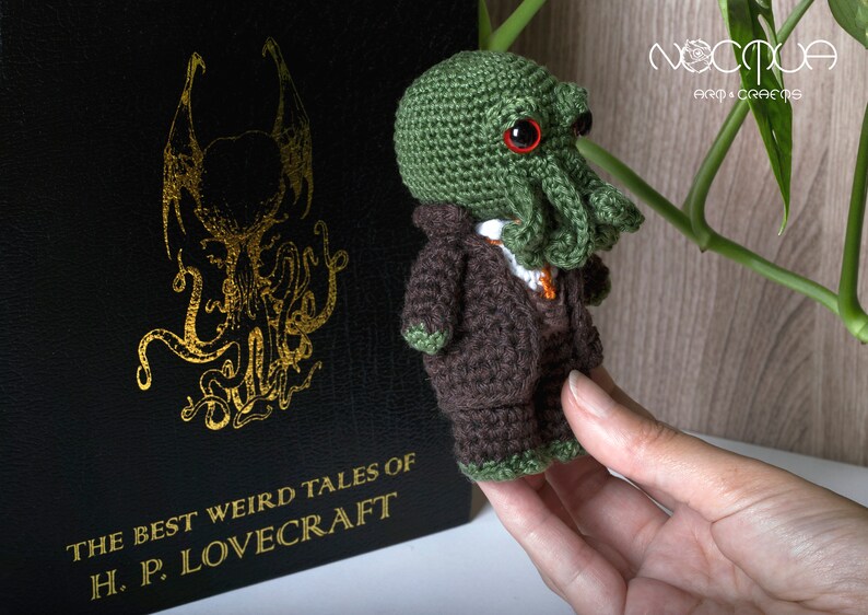 Pode incluir: Um brinquedo de pel&uacute;cia verde e marrom de croch&ecirc; em forma de monstro Cthulhu vestindo um terno marrom. O brinquedo est&aacute; sendo segurado em uma m&atilde;o. O fundo &eacute; um livro com o t&iacute;tulo "The Best Weird Tales of H.P. Lovecraft".