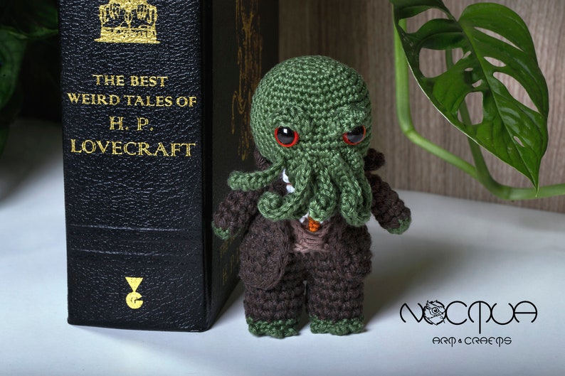 Pode incluir: Um brinquedo de pel&uacute;cia Cthulhu verde de croch&ecirc; vestindo um terno marrom. O brinquedo est&aacute; em p&eacute; ao lado de um livro intitulado "The Best Weird Tales of H.P. Lovecraft".
