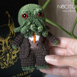 Pode incluir: Um brinquedo de pel&uacute;cia de croch&ecirc; de Cthulhu vestindo um terno marrom e uma gravata verde. O brinquedo tem olhos vermelhos e est&aacute; segurando um pequeno objeto laranja na m&atilde;o.
