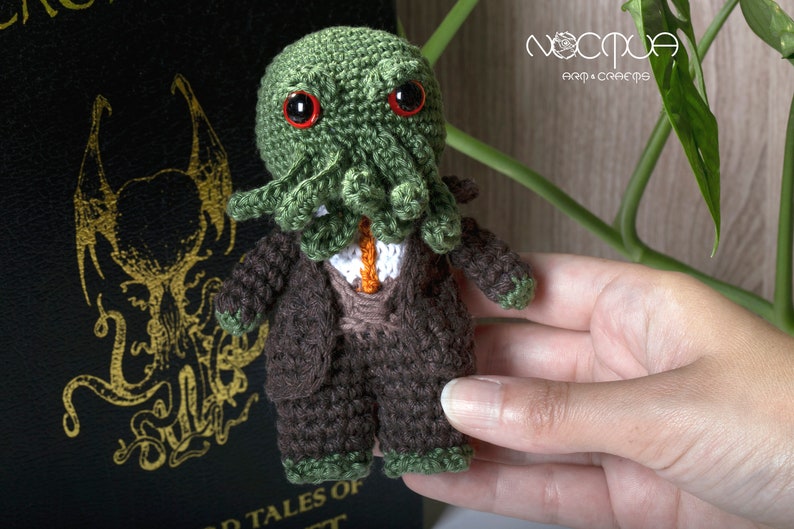 Pode incluir: Um brinquedo de pel&uacute;cia de croch&ecirc; de Cthulhu vestindo um terno marrom e uma gravata verde. O brinquedo tem olhos vermelhos e est&aacute; segurando um pequeno objeto laranja na m&atilde;o.