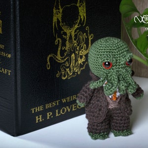 Pode incluir: Um brinquedo de pel&uacute;cia Cthulhu de croch&ecirc; verde e marrom em p&eacute; na frente de um livro intitulado "The Best Weird Tales of H.P. Lovecraft".