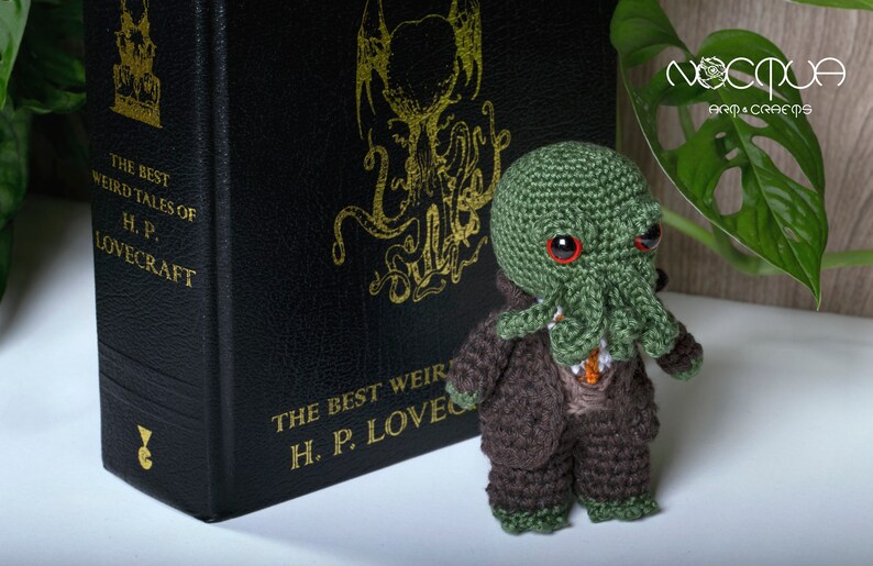 Pode incluir: Um brinquedo de pel&uacute;cia Cthulhu de croch&ecirc; verde e marrom em p&eacute; na frente de um livro intitulado "The Best Weird Tales of H.P. Lovecraft".