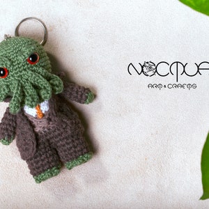 Pode incluir: Chaveiro Cthulhu de croch&ecirc; verde com olhos vermelhos, vestindo um terno marrom e uma gravata. O chaveiro est&aacute; preso a um anel prateado. O fundo &eacute; branco com uma folha verde.