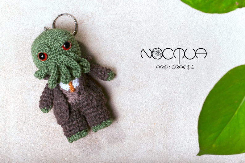Pode incluir: Chaveiro Cthulhu de croch&ecirc; verde com olhos vermelhos, vestindo um terno marrom e uma gravata. O chaveiro est&aacute; preso a um anel prateado. O fundo &eacute; branco com uma folha verde.
