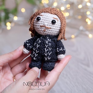 Könnte beinhalten: Gehäkelte Amigurumi-Puppe mit braunen Haaren, einem schwarz-weiß gemusterten Pullover und einem Schnurrbart. Die Puppe hält ein kleines, weißes Objekt in der Hand.