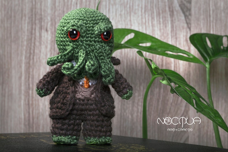 Pode incluir: Um brinquedo de pel&uacute;cia verde de croch&ecirc; com uma cabe&ccedil;a de tent&aacute;culos e olhos vermelhos, vestindo um terno marrom. O brinquedo est&aacute; em p&eacute; sobre uma superf&iacute;cie escura com uma planta verde no fundo. O texto "NOCCTUA ART & CRAFTS" &eacute; vis&iacute;vel no fundo.
